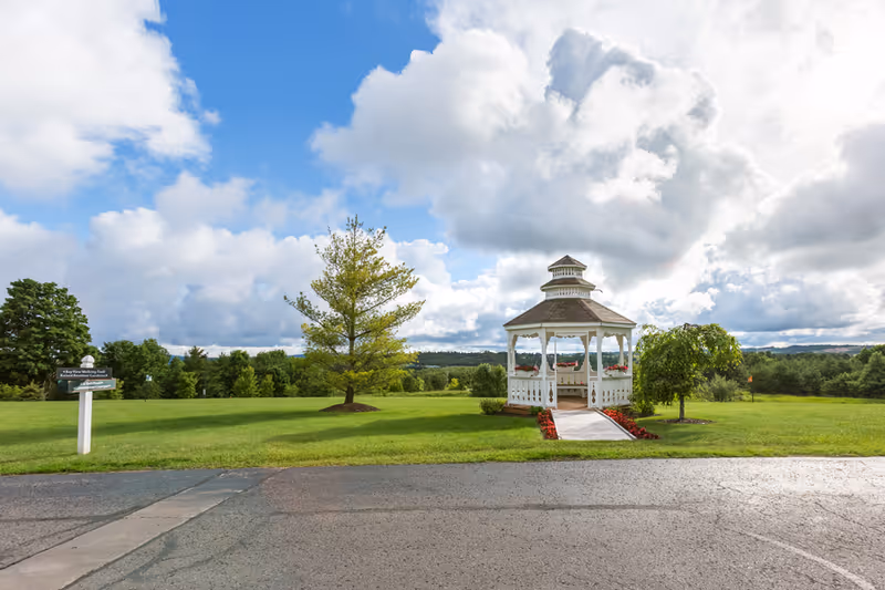 A white gazebo with a shingled roof sits on a well-maintained grassy area with a paved path leading to it. The gazebo is surrounded by red flowers and small trees, with a partly cloudy blue sky overhead.