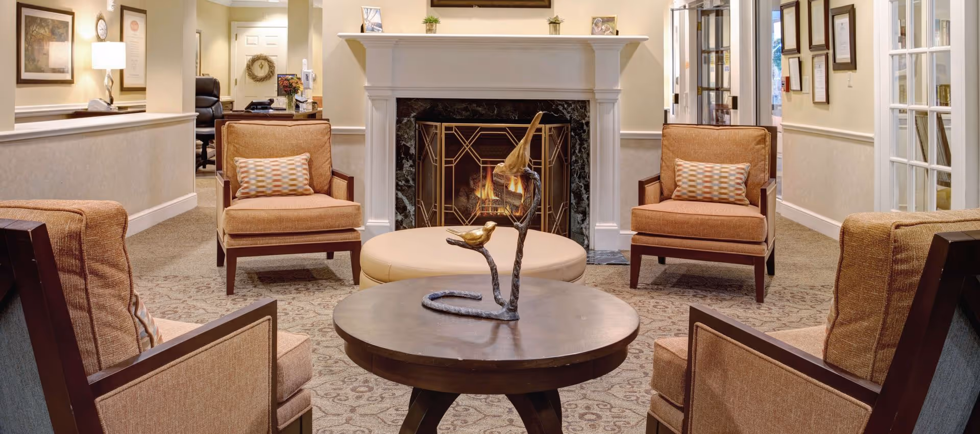 A cozy living room area with four cushioned armchairs arranged around a round wooden coffee table with a decorative bird sculpture. Behind the table is a round ottoman and a fireplace with a fire burning inside. The room has beige walls, carpeted floor, framed pictures on the walls, and a desk with a chair in the background.