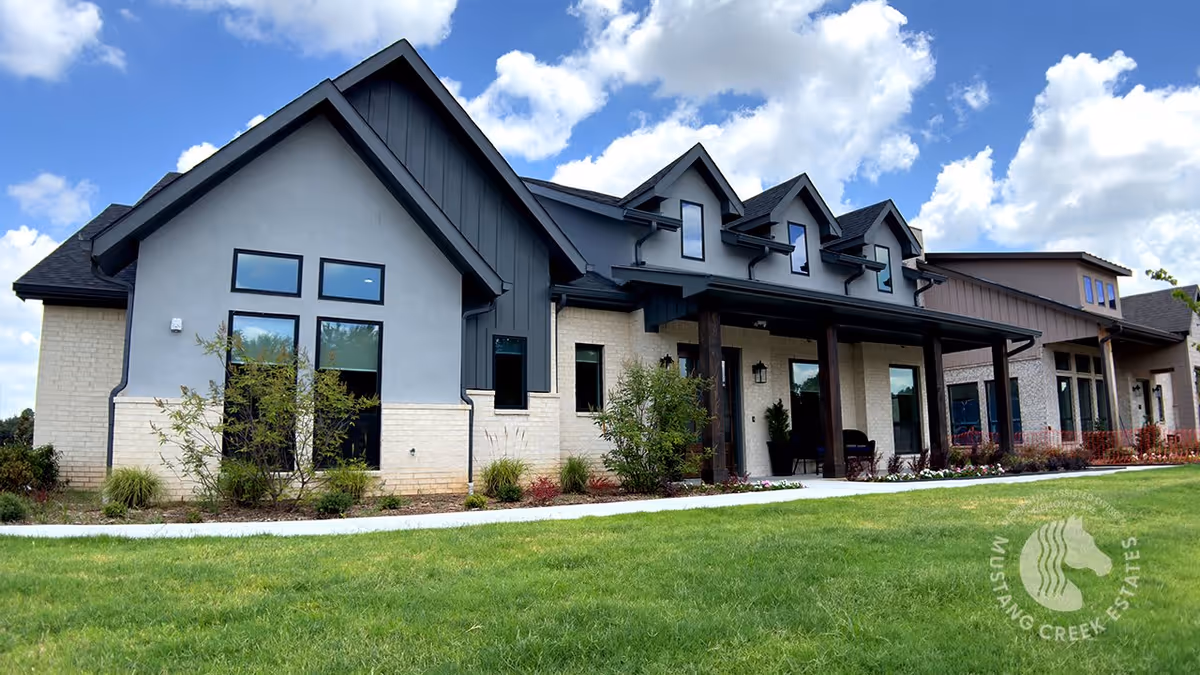 Exterior view of a modern senior living facility building with a combination of light brick and dark panel siding, multiple windows, and a covered porch area. The building is surrounded by a well-maintained lawn and landscaping under a partly cloudy sky.