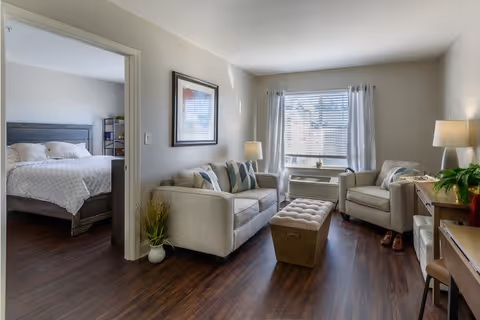A cozy living room with a beige sofa, an armchair, a cushioned ottoman, a side table with a lamp, and a window with white curtains letting in natural light. Adjacent to the living room is a bedroom with a bed, visible through an open doorway.