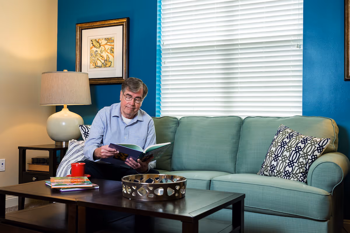 An elderly man sitting on a light green couch in a living room with blue walls, reading a book. There is a wooden coffee table in front of him with a red mug, some books, and a decorative metal tray. A table lamp and framed artwork are visible on a side table next to the couch.