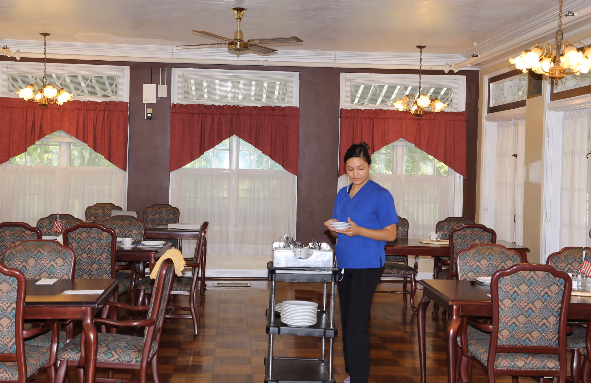 A dining room with multiple wooden tables and patterned upholstered chairs. The room has large windows with red curtains and white sheer drapes. A woman in a blue uniform is standing near a black serving cart holding a stack of small plates. The ceiling has ceiling fans and hanging light fixtures.