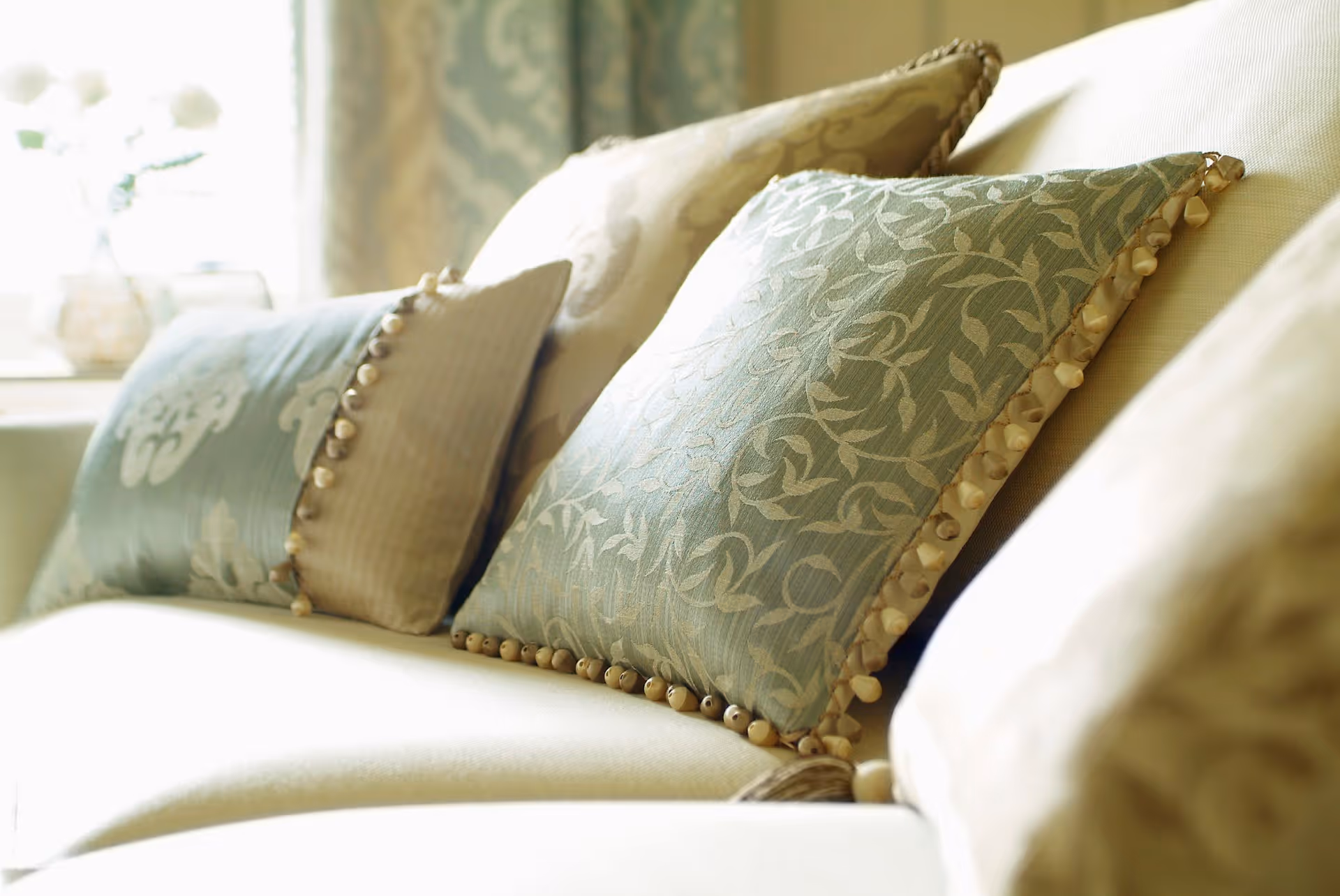 Close-up view of decorative cushions with intricate patterns and tassels arranged on a light-colored sofa in a softly lit room.