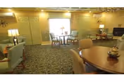 A cozy living room area with multiple seating arrangements including armchairs and round tables. The room features carpeted flooring, soft yellow walls, and two lamps providing warm lighting. There is a window with blinds letting in natural light, and decorative wall art is visible. The space appears comfortable and inviting.