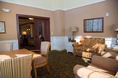 A cozy living room area with a patterned loveseat, two armchairs, a white side table with a lamp, and a framed painting on the wall. There is a wooden table with striped upholstered chairs in the foreground and double wooden doors leading to another room in the background.