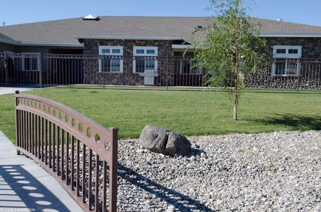 Outdoor view of a senior living facility with a stone and brick building in the background, a small tree, a grassy lawn, a rock garden, and a brown metal railing along a concrete pathway.
