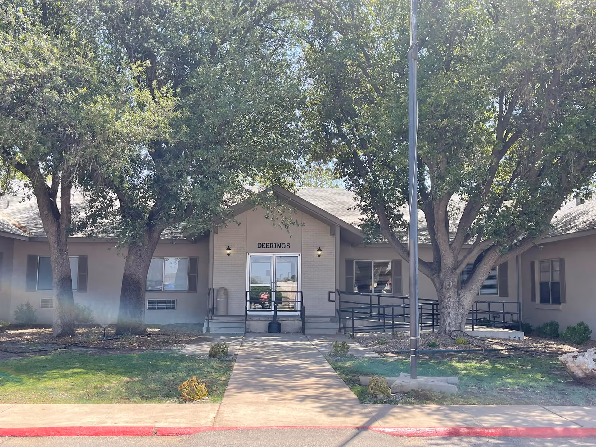Front exterior view of Deerings Nursing and Rehabilitation building with a walkway leading to double glass doors. The building is surrounded by large trees and has a wheelchair ramp on the right side.