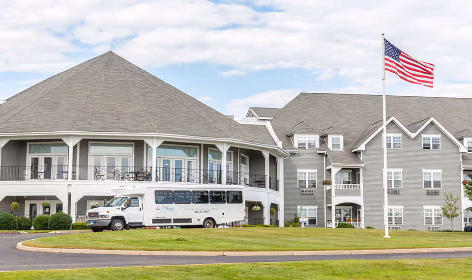Front exterior of a gray senior living facility with a shuttle bus parked in front and an American flag on a pole.