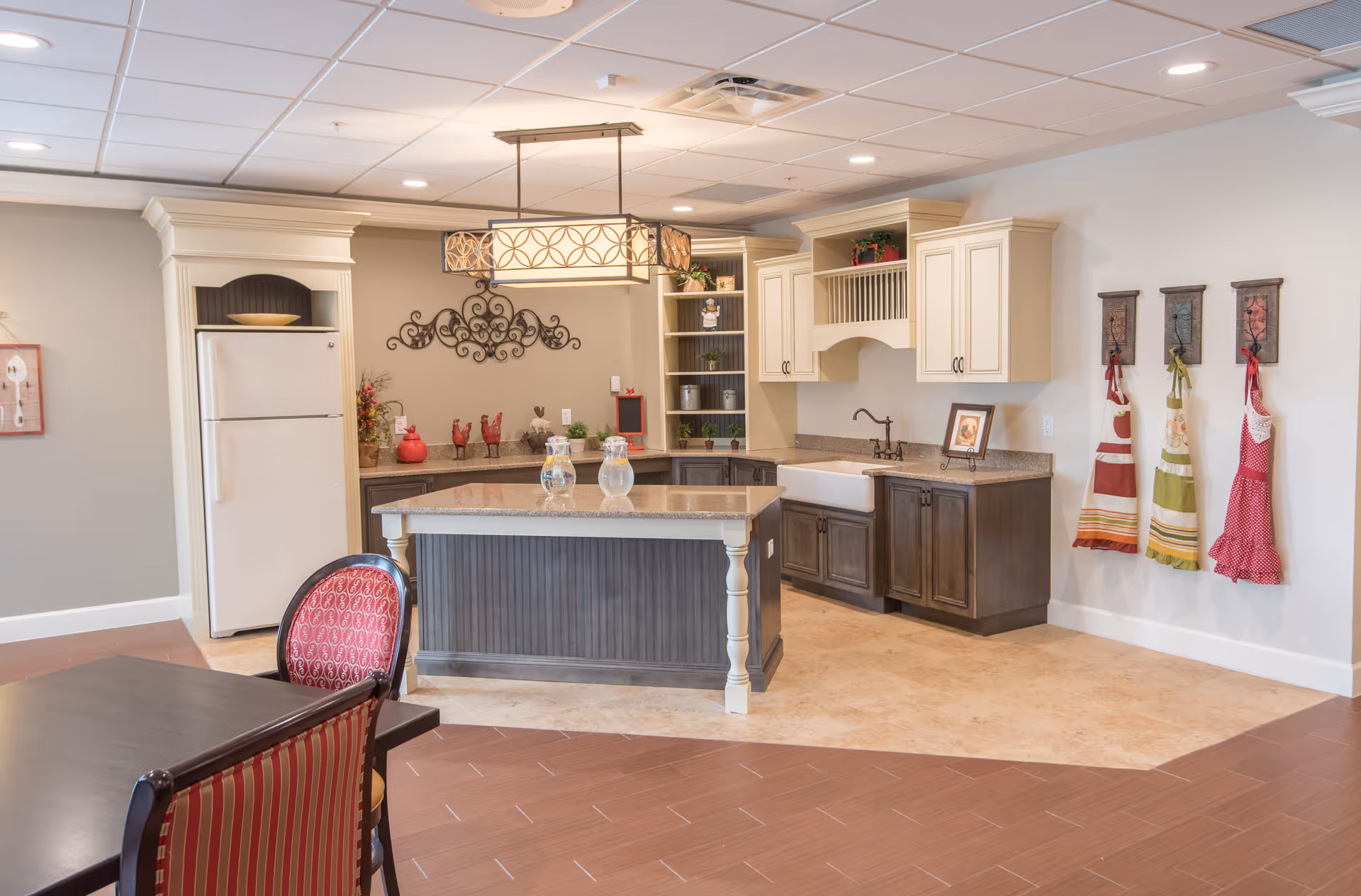 A bright and spacious kitchen area with a central island featuring a granite countertop. The kitchen has cream-colored upper cabinets and dark lower cabinets, a white farmhouse sink, and a white refrigerator. Three colorful aprons hang on the wall to the right, and a dining table with red and gold patterned chairs is partially visible in the foreground. The ceiling has recessed lighting and a decorative hanging light fixture above the island.