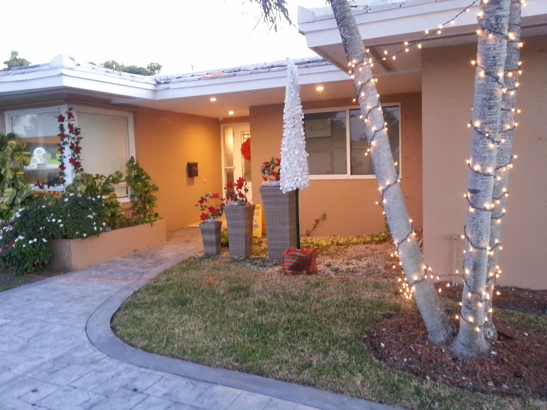 Entrance of a single-story building with a covered porch area, decorated with string lights wrapped around two palm trees and holiday decorations including a white cone-shaped ornament and red flowers in tall planters. The walkway leading to the entrance is paved and bordered by grass and plants.