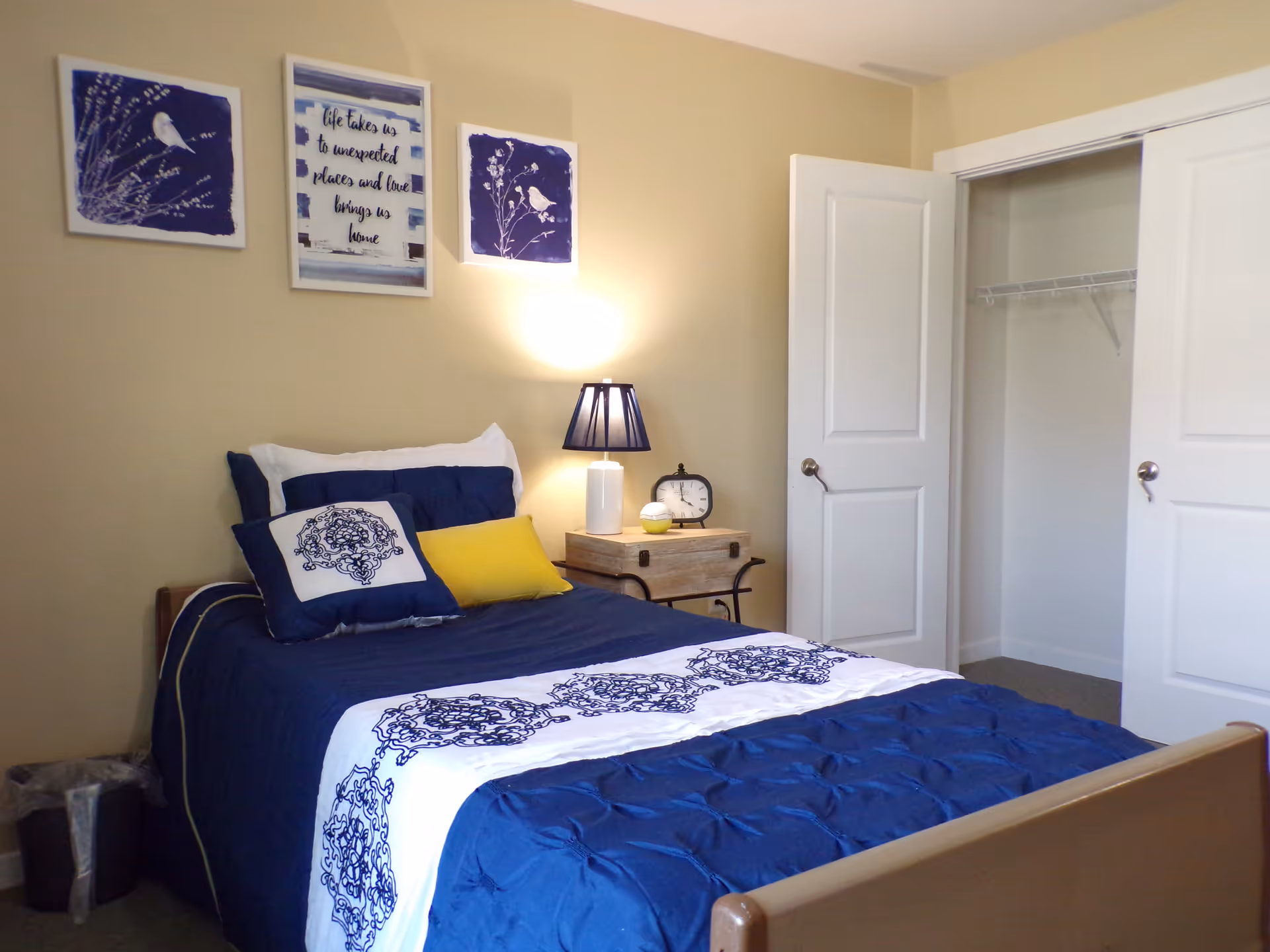 A bedroom with a single bed made with blue and white bedding, accented by a yellow pillow. Above the bed are three framed pictures, two with bird designs and one with a quote. A nightstand beside the bed holds a lamp and a clock. There is an open closet with double doors in the background.