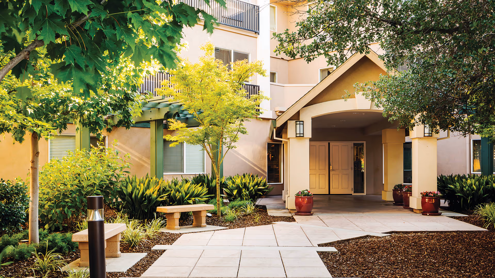 Outdoor entrance area of Atria Carmichael Oaks facility with a paved walkway, benches, green trees, shrubs, and potted plants near the building entrance under a covered porch.