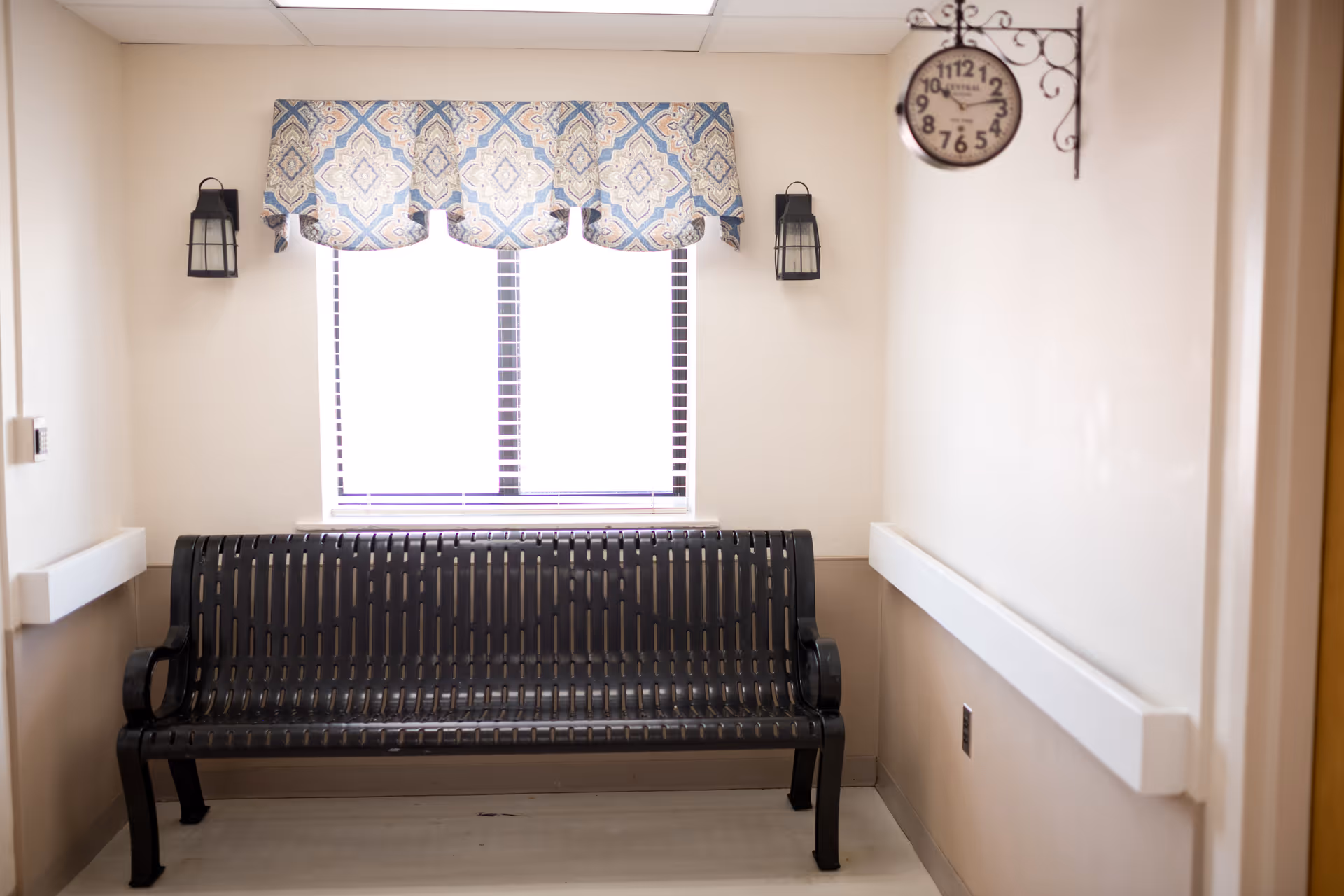 A small waiting area with a black metal bench placed against a wall beneath a window with a patterned valance. Two black wall-mounted lantern-style lights flank the window. A decorative clock with Roman numerals hangs on the right wall. The walls are painted in a light beige color with white handrails on both sides.