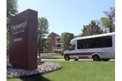 Outdoor view of a senior living facility with a brown sign reading 'Parkwood Senior Living Entrance' on a grassy area with rocks. A white shuttle van is parked on the driveway near the building, surrounded by trees and clear blue sky.