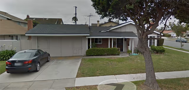 Single-story residential building with a driveway and a parked black car in front. The house has a garage, a small front porch with bushes, and a large tree on the lawn near the sidewalk.