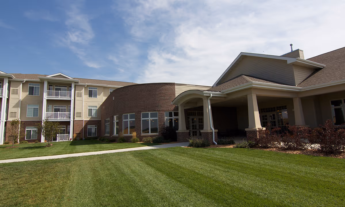 Exterior view of Whispering Creek Senior Living facility showing a multi-story building with beige siding and brick accents, large windows, balconies, a covered entrance, and a well-maintained green lawn under a partly cloudy sky.