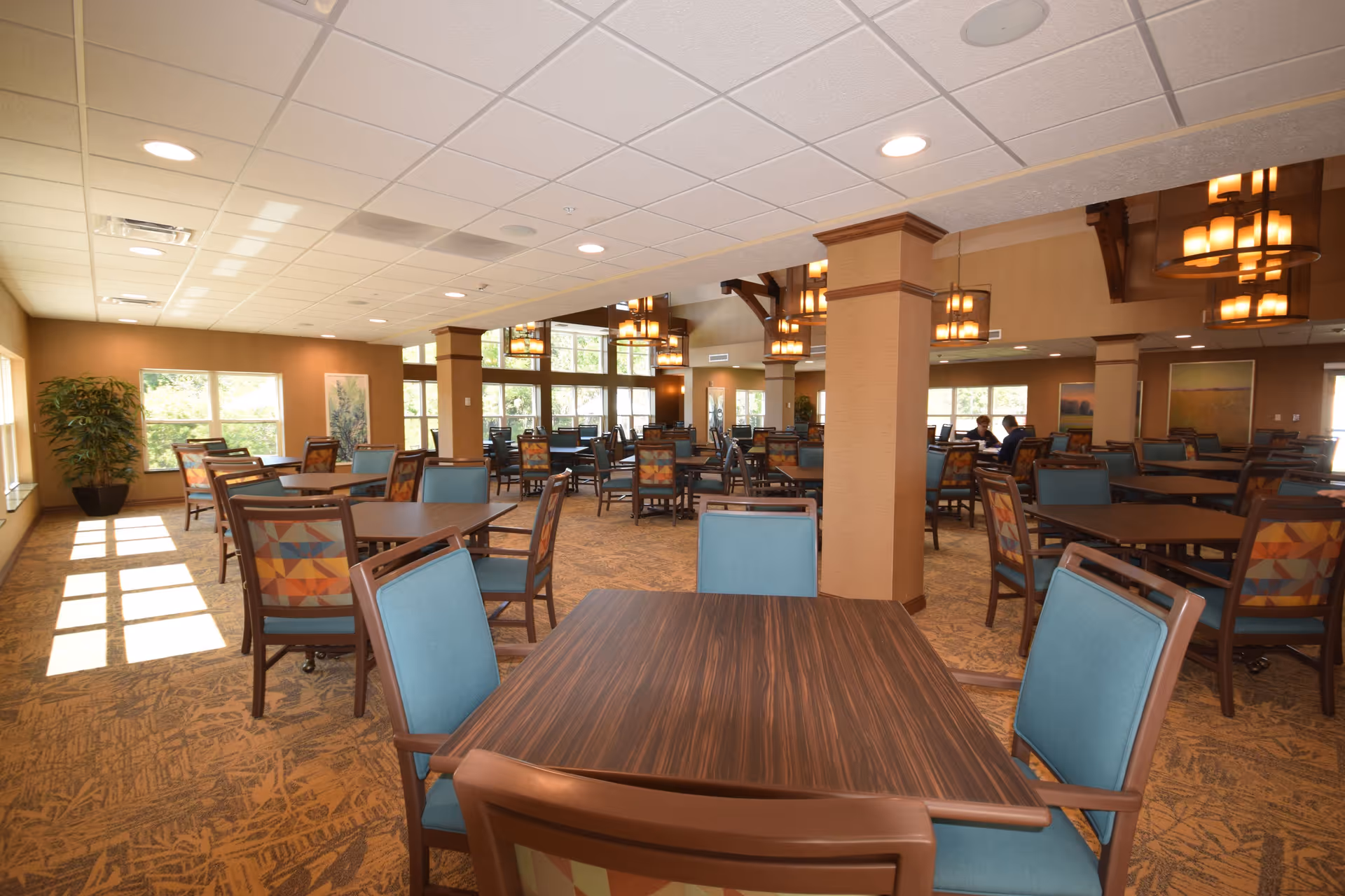 A spacious dining room in a retirement community with multiple wooden tables and chairs featuring blue and patterned upholstery. Large windows allow natural light to fill the room, and warm pendant lights hang from the ceiling. The room has a carpeted floor and beige walls with decorative columns.