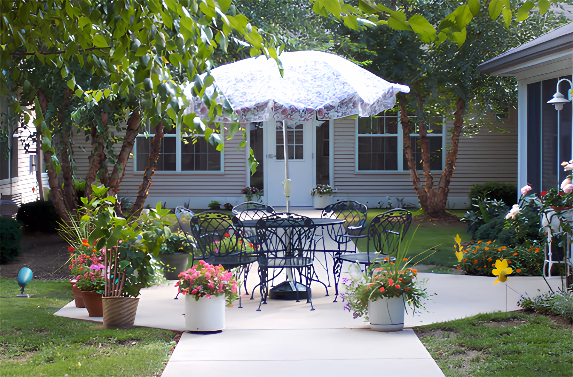 A shaded outdoor patio with a round metal table and chairs under a patterned umbrella surrounded by potted flowers in front of a residential building.