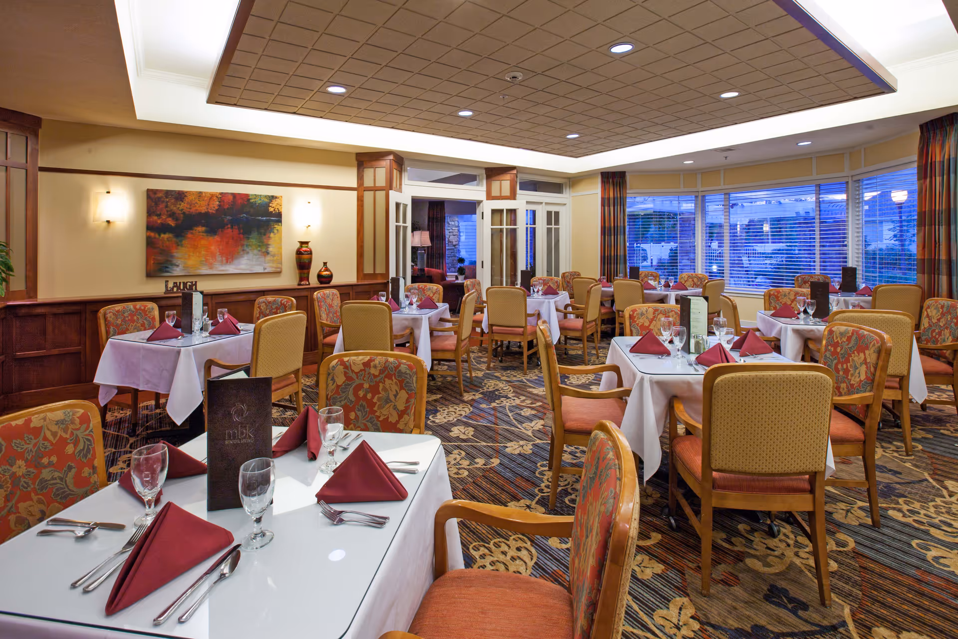 A well-lit dining room with multiple tables covered in white tablecloths, each set with burgundy folded napkins, glassware, and silverware. The room features patterned carpet, floral upholstered chairs, large windows with blinds, and warm wall lighting. A colorful painting and decorative vases are visible on the wall.