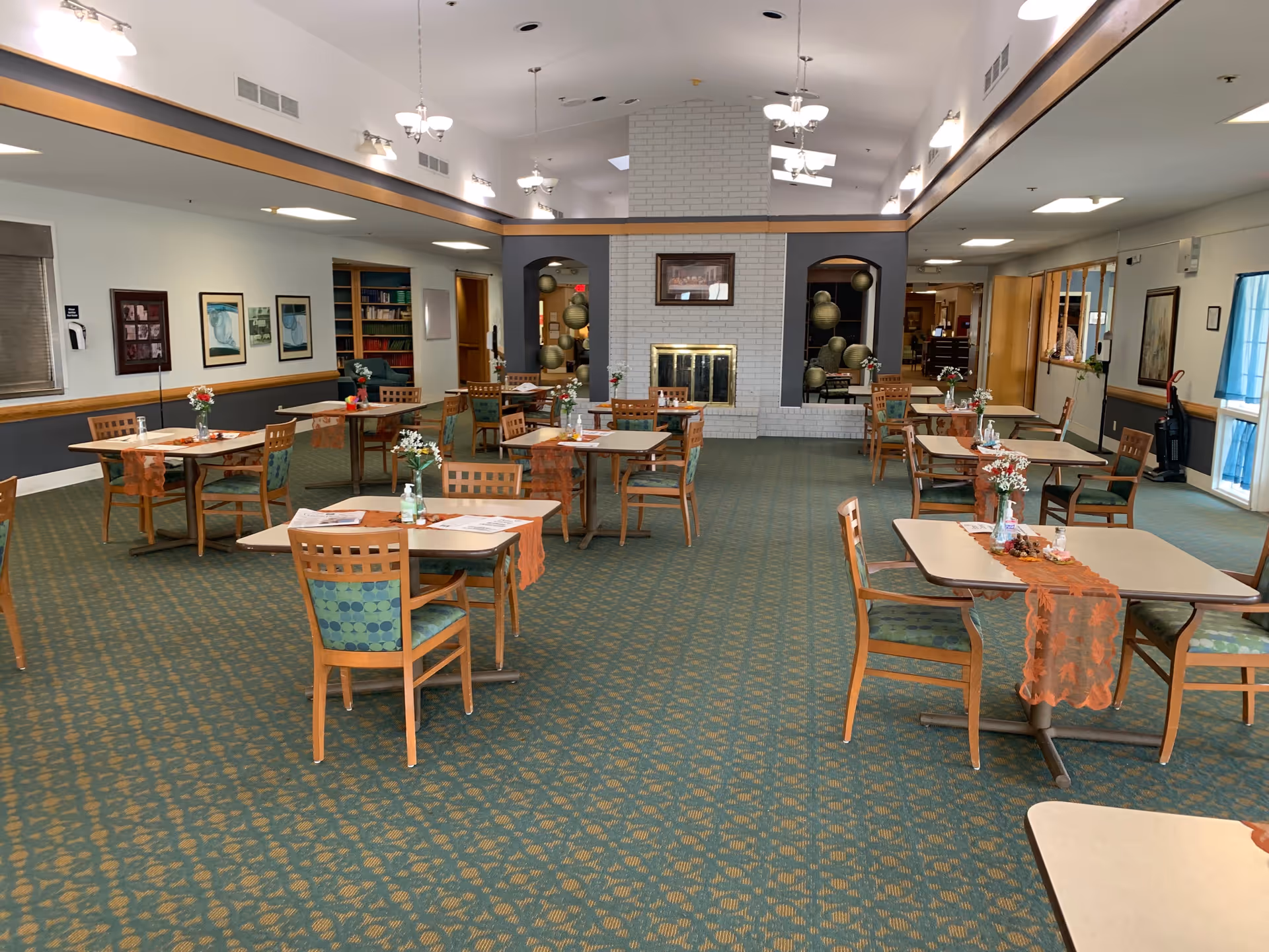 A spacious dining room with multiple tables and chairs arranged neatly. Each table has a small vase with flowers and an orange table runner. The room features a green patterned carpet, a white brick fireplace in the center, and large mirrors on the walls. There are several light fixtures hanging from the ceiling and framed pictures on the walls.