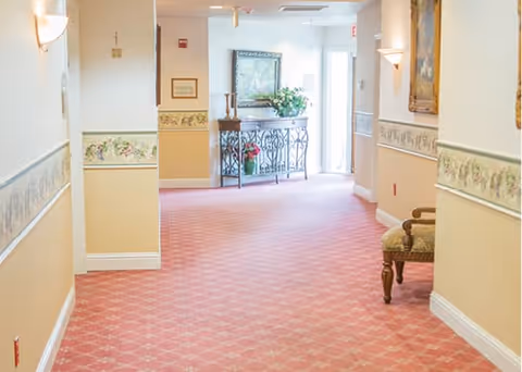 A well-lit hallway in an assisted living facility with patterned pink carpet, beige and white walls with floral border wallpaper, wall sconces, framed artwork, a decorative table with flowers and candles at the end, and a cushioned chair on the right side.