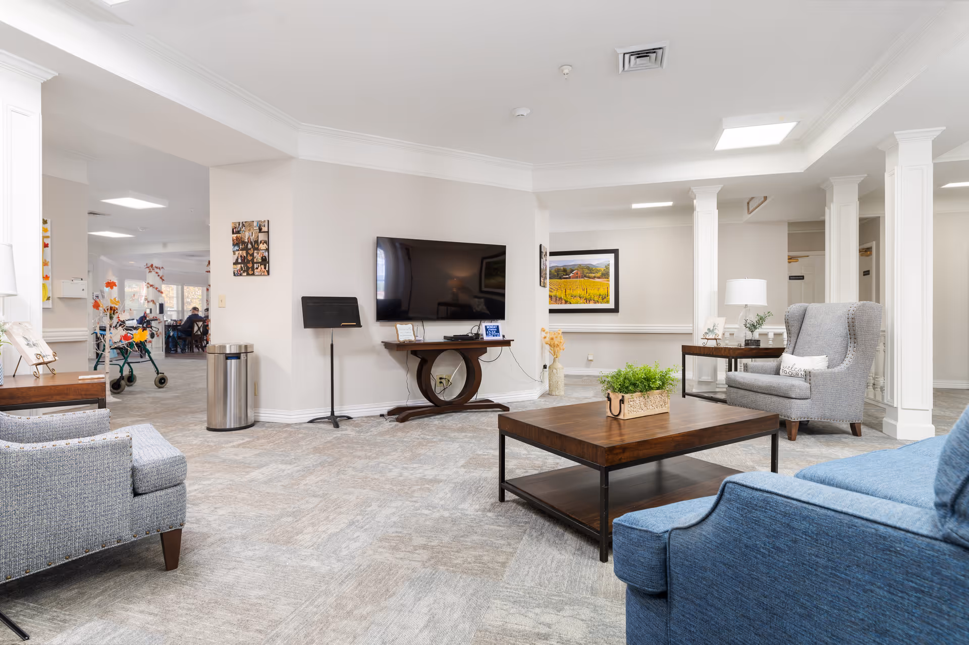 A bright and spacious senior living facility common area with comfortable seating including a blue armchair, a gray wingback chair, and a gray sofa. A wooden coffee table with a green plant centerpiece is in the middle. A flat-screen TV is mounted on the wall above a decorative wooden console table. The room has light-colored walls, carpeted floors, white columns, and framed artwork on the walls. In the background, there is a glimpse of a dining area with people seated and a walker near the entrance.