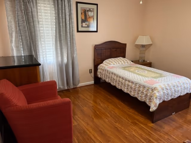 Small furnished bedroom with a single bed, red armchair, wooden dresser, bedside table with lamp, and a curtained window.
