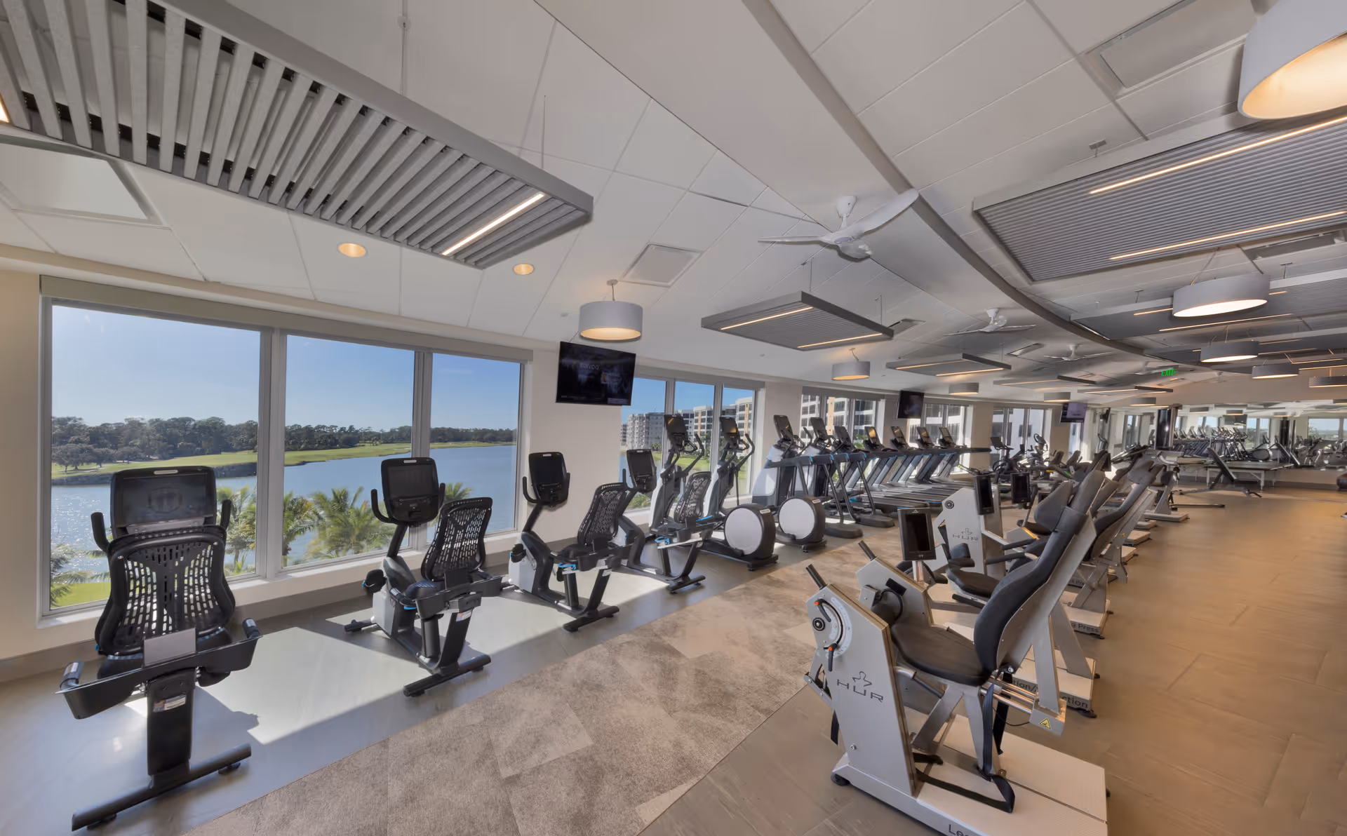 A spacious, modern gym with multiple exercise bikes and treadmills arranged in rows. Large windows provide a scenic view of a lake and greenery outside. The ceiling has contemporary lighting fixtures and fans, and there are several televisions mounted on the walls.