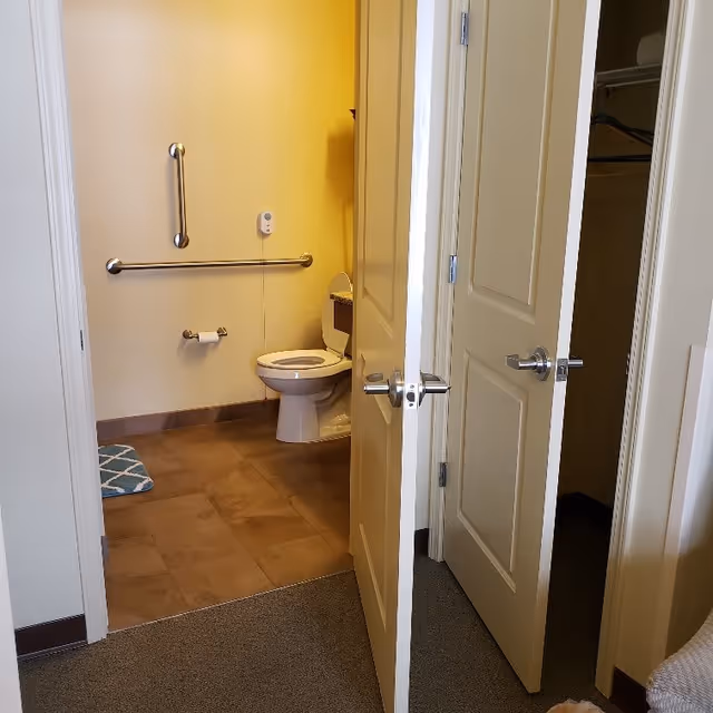 View through an open door into a bathroom with a toilet, grab bars on the wall, a toilet paper holder, and a blue patterned bath mat on the floor. Adjacent to the bathroom door is another door leading to a closet with hangers inside.