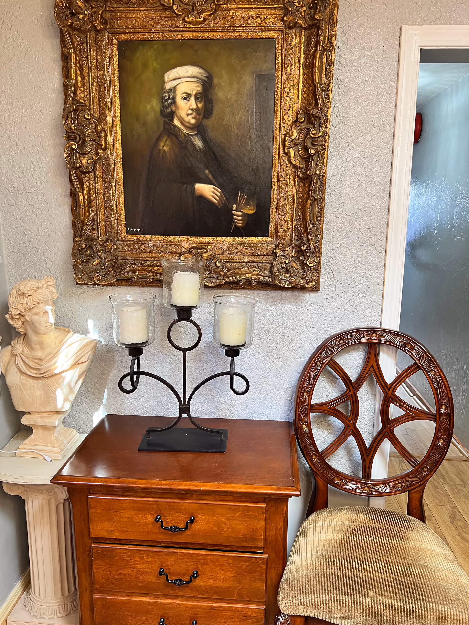 A corner of a room featuring a wooden chest of drawers with a black metal candle holder holding three white candles on top. To the left is a white bust sculpture on a pedestal, and above the chest hangs a framed classical portrait of a man in historical attire. To the right of the chest is a wooden chair with an ornate circular backrest and a cushioned seat. The walls are textured and painted light gray.