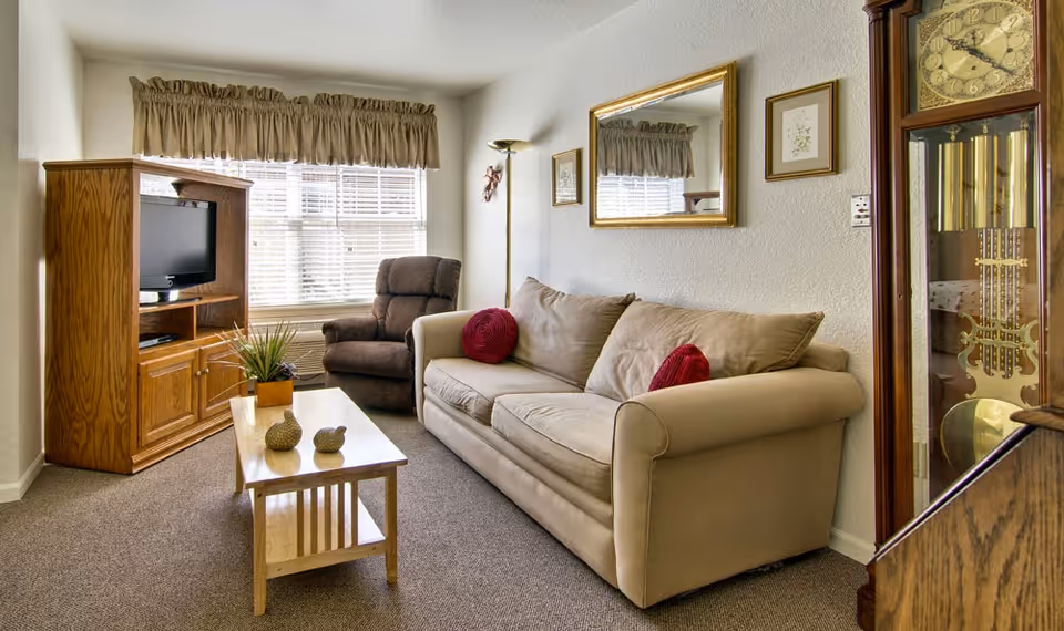 A cozy living room with a beige sofa adorned with two red round pillows, a brown recliner chair, a wooden coffee table with decorative items, a wooden TV cabinet with a television, a tall grandfather clock, and a large window with beige curtains allowing natural light to fill the room.