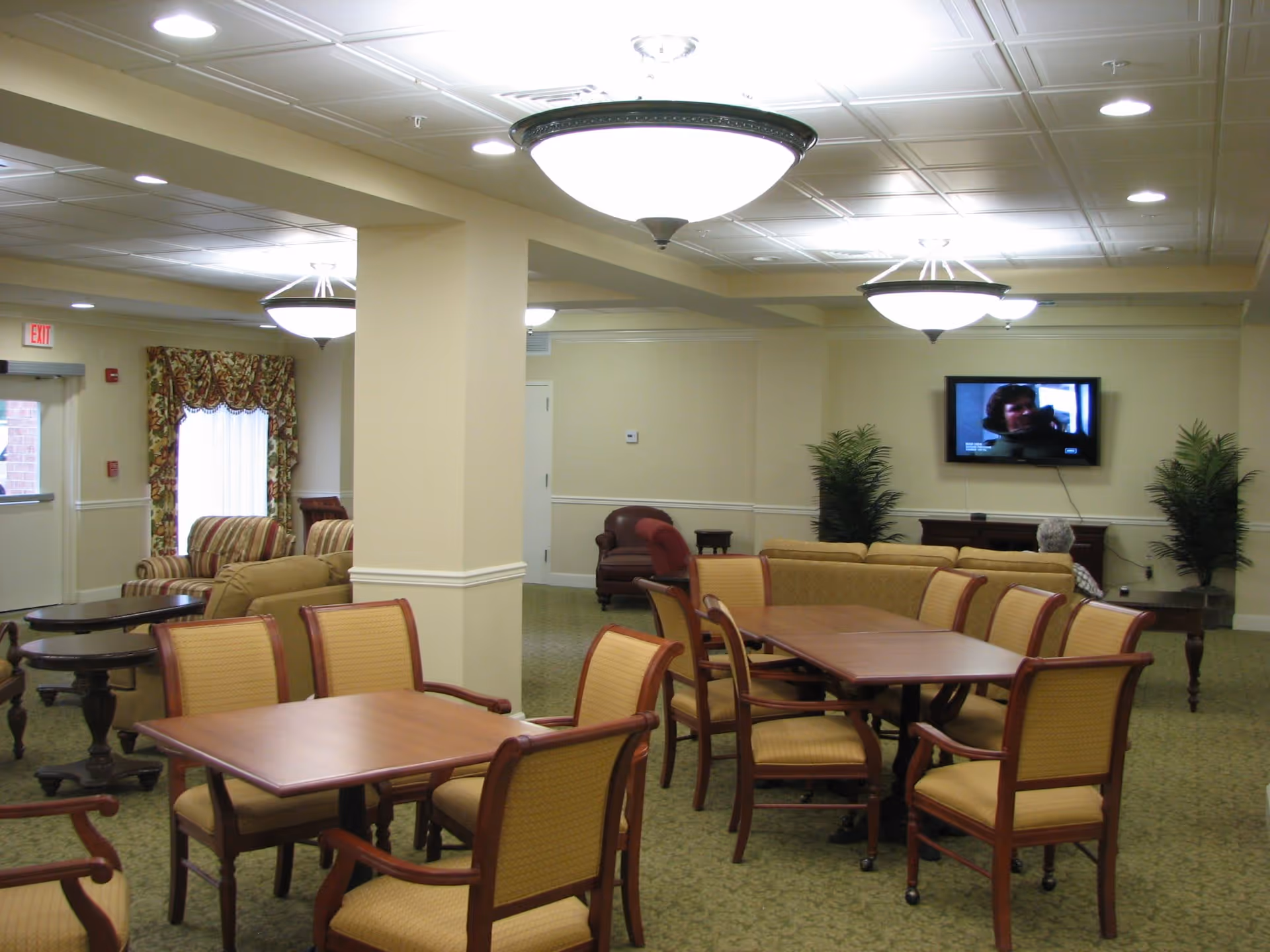 Spacious senior living common area with dining tables and chairs, lounge seating, plants, and a wall-mounted TV.