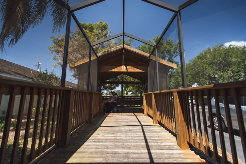 A wooden ramp with railings on both sides leads to a covered seating area with a wooden roof. The structure is enclosed with mesh screens. Trees and a clear blue sky are visible in the background, along with a parked silver car to the right.