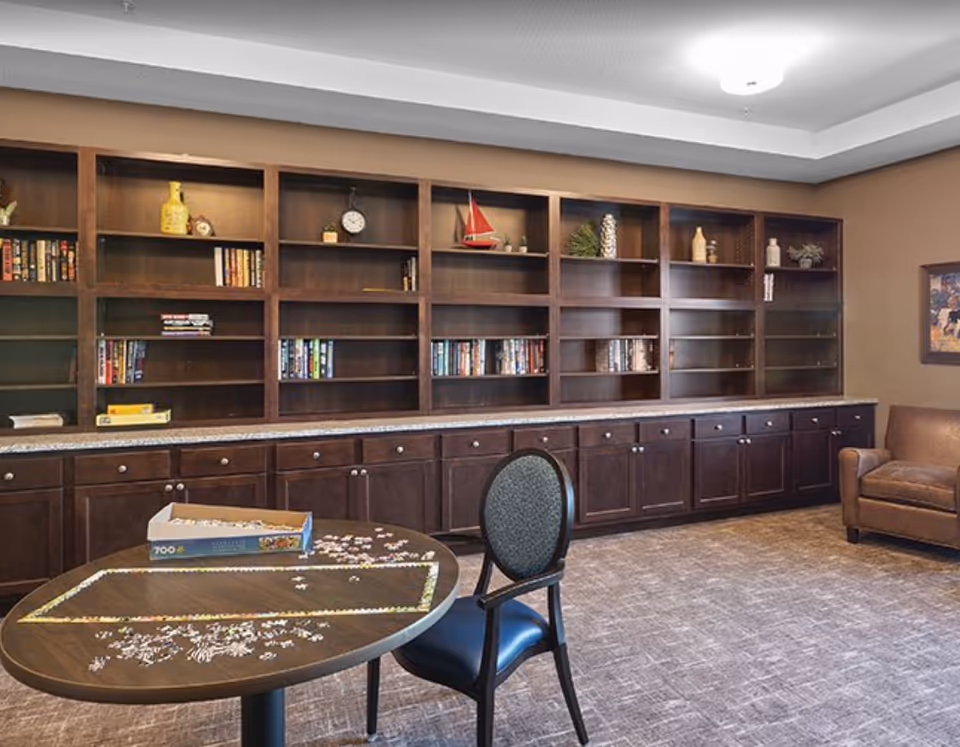 A cozy room with a large wooden bookshelf filled with books and decorative items, a round table with a partially completed jigsaw puzzle and a box of puzzle pieces, a single chair with a cushioned seat, and a brown leather armchair in the corner. The room has beige walls and carpeted flooring.