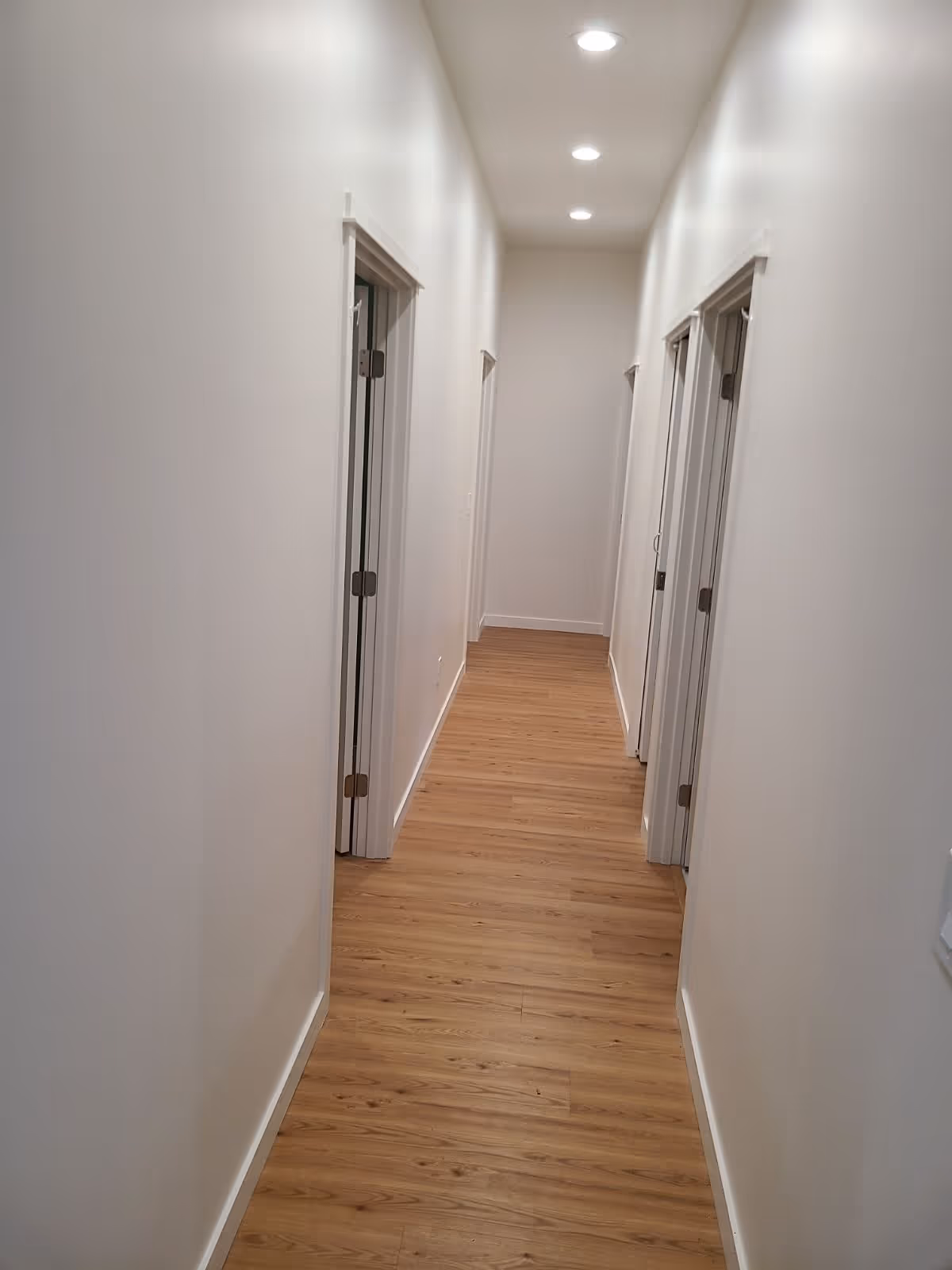 A long, narrow hallway with light wooden flooring and white walls. There are four doorways visible, two on each side of the hallway, and recessed ceiling lights evenly spaced along the ceiling.