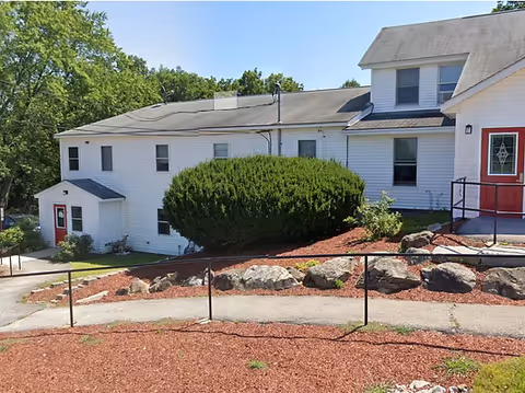 Exterior view of Rose Haven Merrimack, a white building with a red front door, manicured shrubs, and rock landscaping.