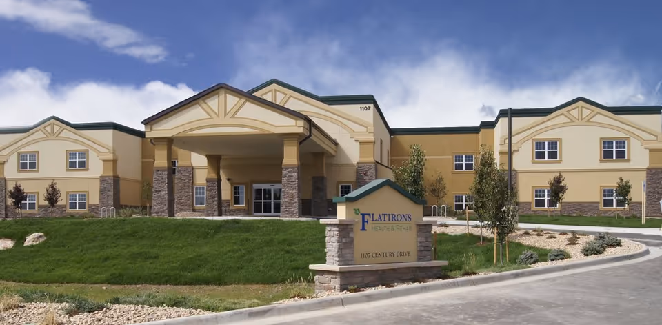 Exterior front view of Flatirons Health & Rehab LLC building with a stone and beige facade, a covered entrance supported by columns, green lawn, small trees, and a sign displaying the facility name and address 1107 Century Drive.