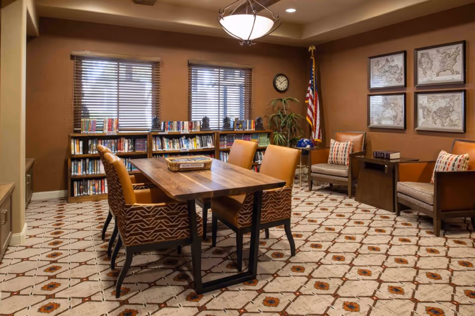 A cozy reading or meeting room with a wooden table surrounded by six patterned chairs. Behind the table, there are bookshelves filled with books against a brown wall with two windows covered by blinds. To the right, there are two armchairs with patterned cushions, a small side table, and four framed maps on the wall. An American flag stands in the corner next to a clock and a potted plant. The floor is covered with a patterned carpet.
