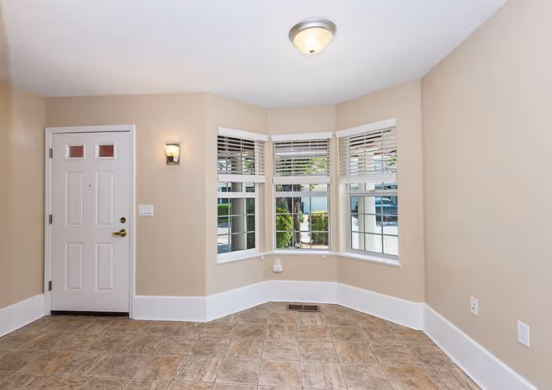 Empty room with beige walls and tiled floor featuring a white front door on the left and a bay window with white blinds overlooking greenery outside. The ceiling has a round light fixture and there is a wall-mounted light next to the door.