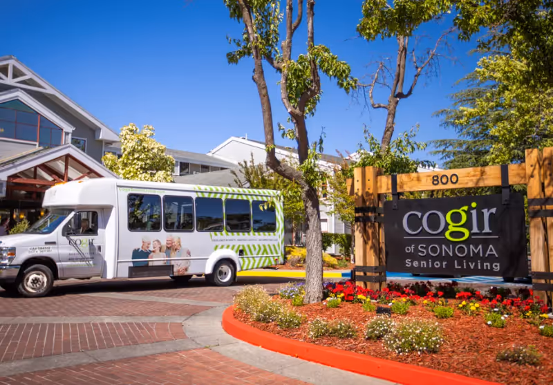 Exterior view of Cogir of Sonoma Senior Living facility with a shuttle bus parked near the entrance. The scene includes a landscaped area with flowers and trees, and a large wooden sign displaying the facility's name and address number 800.