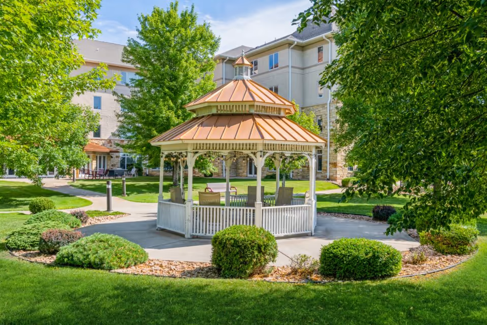 A white gazebo with a copper-colored roof sits in the center of a landscaped garden area with green grass, bushes, and trees. Behind the gazebo is a multi-story senior living facility building with beige and stone exterior walls and several windows. The sky is clear and blue.