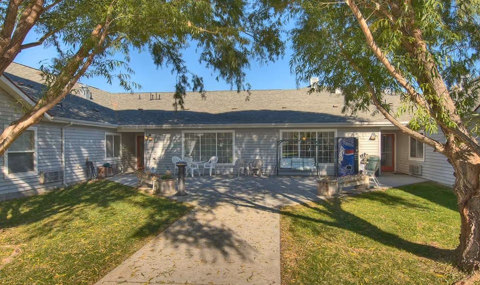 Outdoor courtyard area of Dorian Place Assisted Living with a paved walkway, green grass, trees providing shade, white chairs and tables, and a vending machine near the building entrance.