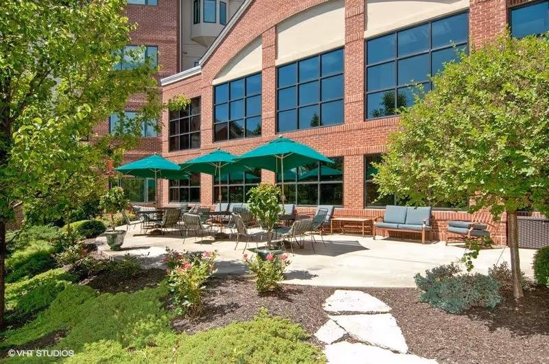 Outdoor patio area at The Wellington at North Bend Crossing with green umbrellas, metal chairs, cushioned benches, surrounded by trees and shrubs in front of a large brick building with many windows.