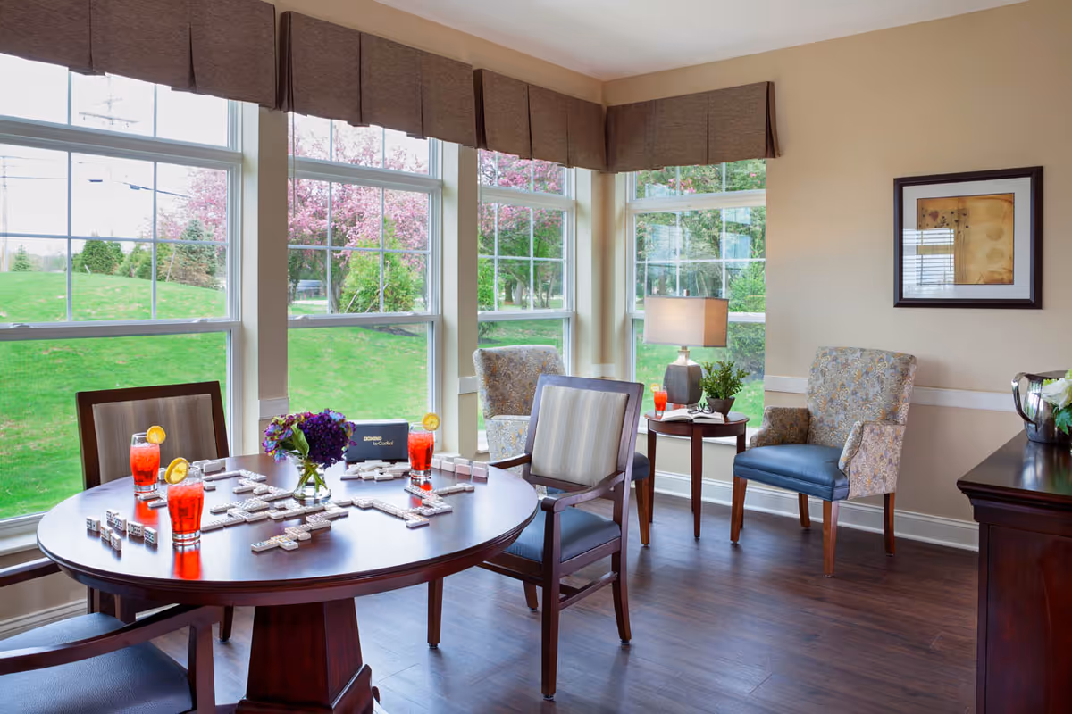 A cozy sitting area with large windows showing a green lawn and blooming trees outside. Inside, there is a round wooden table with dominoes set up for a game and three glasses of red drink garnished with lemon slices. Around the table are wooden chairs with cushions. In the corner, there are two upholstered armchairs next to a small wooden side table with a lamp, a plant, and another glass of red drink. A framed artwork hangs on the beige wall.