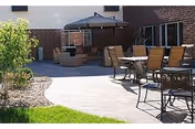 Outdoor patio area with several tables and chairs arranged on a paved surface next to a building. There is a seating area with cushioned furniture under a canopy, surrounded by some landscaping including grass and a small tree.