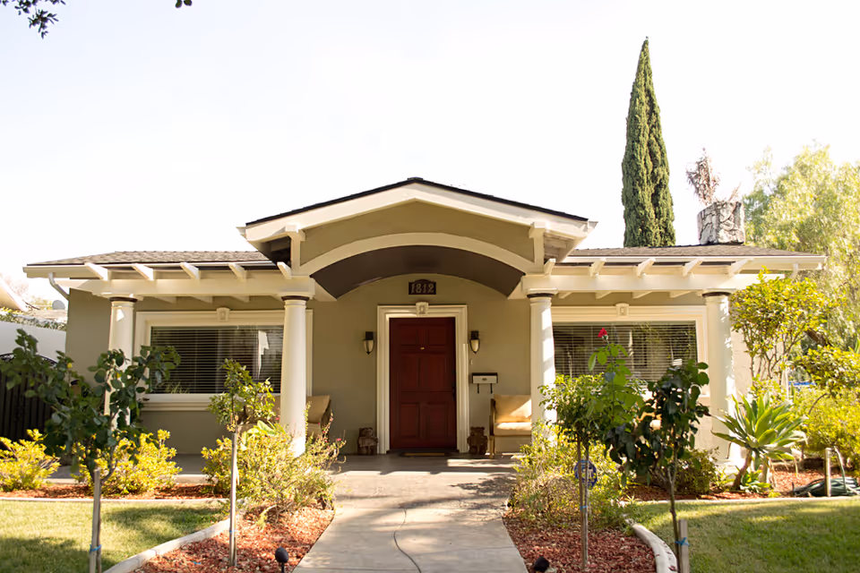 Front exterior view of a single-story residential care facility building with a covered porch, two white columns, a red door, large windows with blinds, and a well-maintained garden with shrubs and small trees.