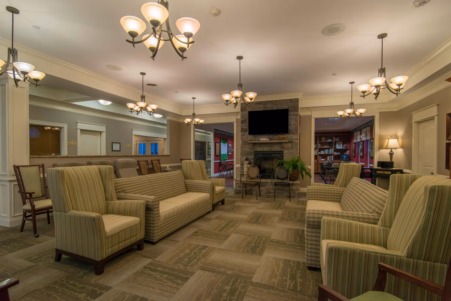 A spacious and well-lit common area with multiple striped armchairs and sofas arranged around a stone fireplace with a mounted flat-screen TV above it. The room features several chandeliers hanging from the ceiling, a patterned carpet floor, and a lamp on a side table. In the background, there are doorways leading to other rooms, including one with bookshelves and a desk.