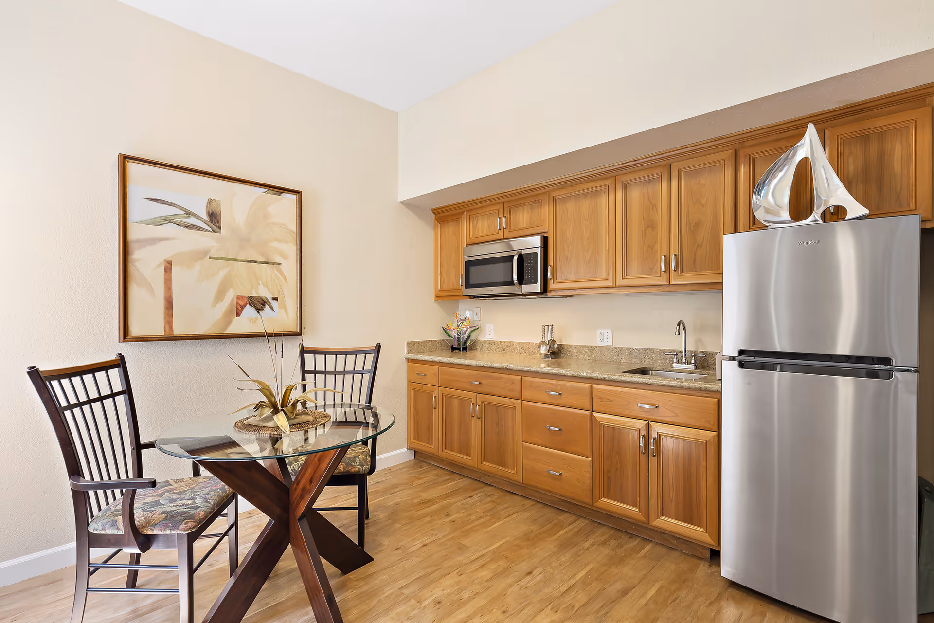A small kitchen area with wooden cabinets, a stainless steel refrigerator, a built-in microwave, and a granite countertop with a sink. In front of the kitchen is a round glass dining table with a wooden base and two chairs with patterned cushions. A framed artwork featuring palm trees hangs on the wall.