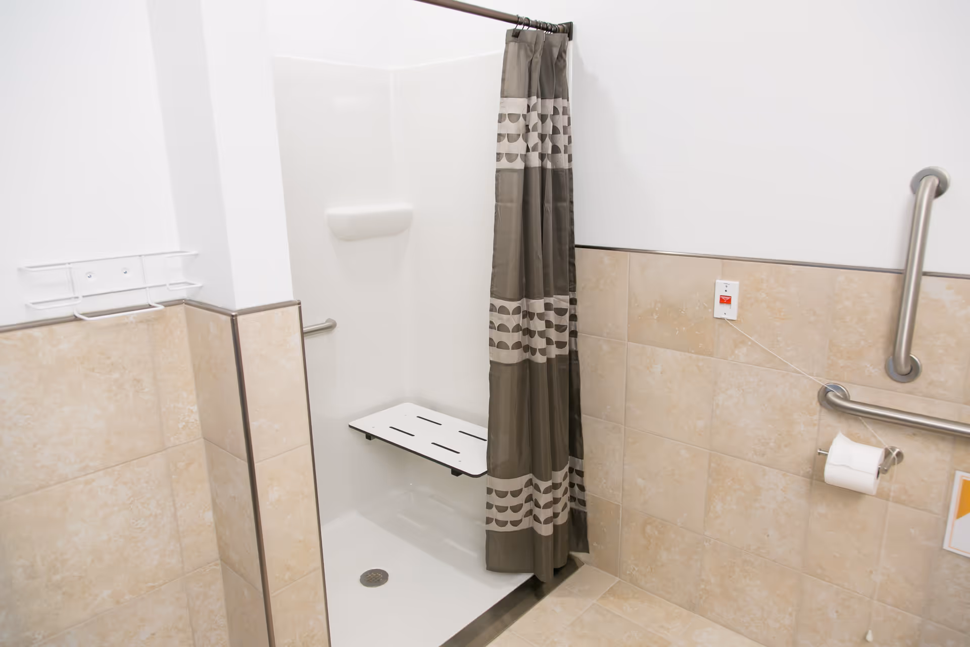 Accessible shower area with a fold-down seat, grab bars, a shower curtain with a geometric pattern, and beige tiled walls. There is an emergency pull cord and a toilet paper holder mounted on the wall.