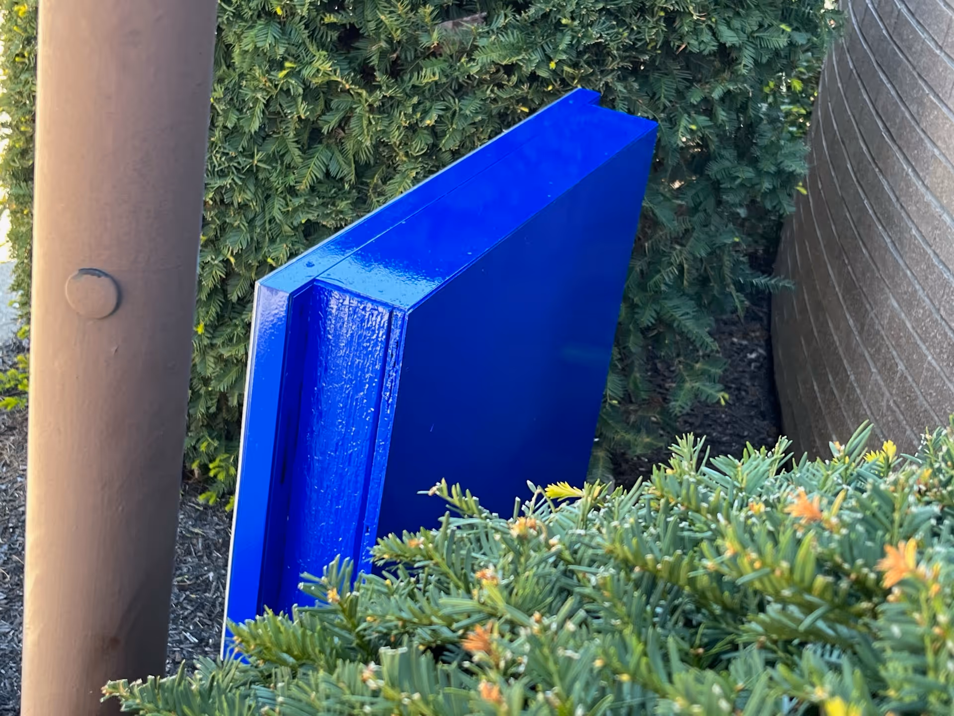 A close-up view of a bright blue rectangular object placed outdoors among green bushes and next to a brown cylindrical pole and a curved brick wall.